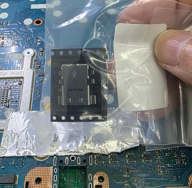Conector de energia novo dentro de um saquinho plástico, segurado pela mão do técnico, com a placa e o papel alumínio ao fundo