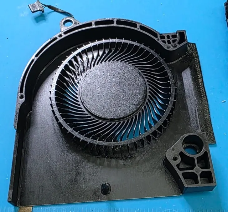 Ventoinha de um Dell G15 5511 fora do notebook, sobre o tapete azul da bancada, com poeira fina acumulada nas pás e na carcaça de plástico