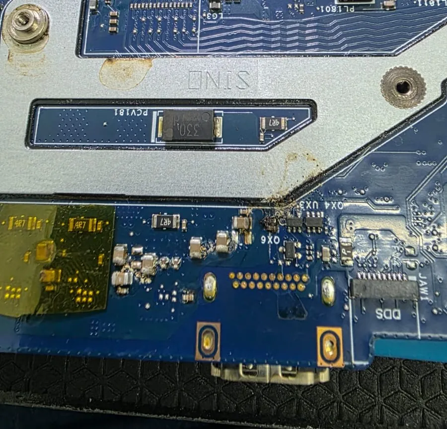 Borda da placa-mãe do Dell G15 5525 com mancha escura de resíduo e pequenos componentes oxidados perto da base metálica