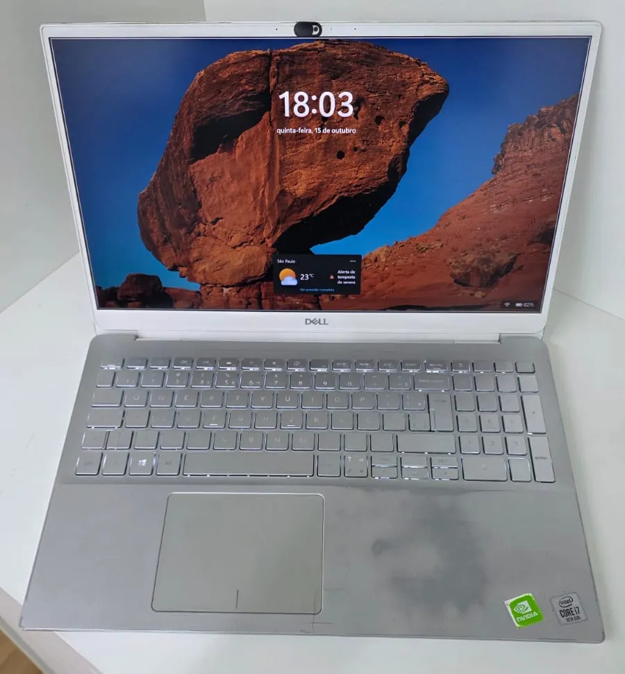 Dell Inspiron 5590 prateado ligado sobre uma bancada branca, com a tela mostrando a área de trabalho do Windows e o teclado com a iluminação acesa
