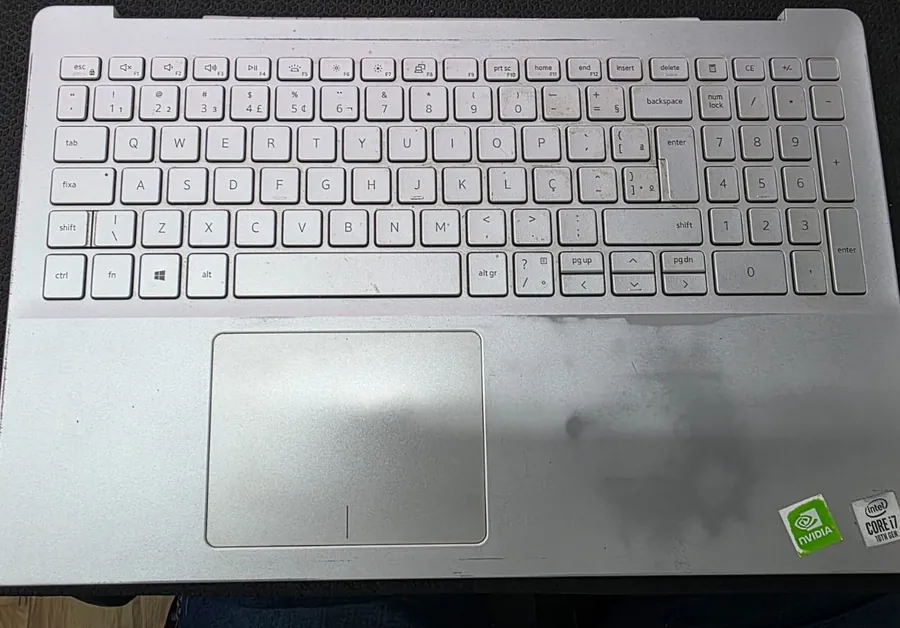 Palmrest de um Dell Inspiron 5590 visto de cima, com o teclado ABNT2 de teclas brancas, o touchpad e manchas escuras no plástico à direita, com os adesivos da NVIDIA e do Core i7 de 10ª geração