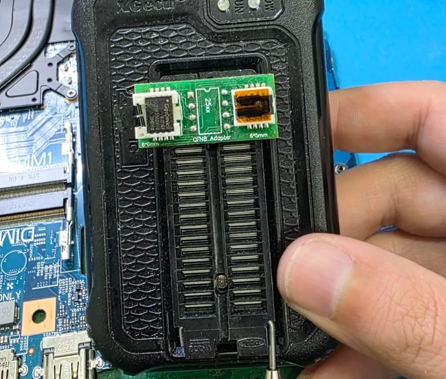 Mão segurando o adaptador do programador XGecu com o chip de BIOS GigaDevice encaixado no soquete de seis por oito milímetros, sobre o soquete ZIF do equipamento