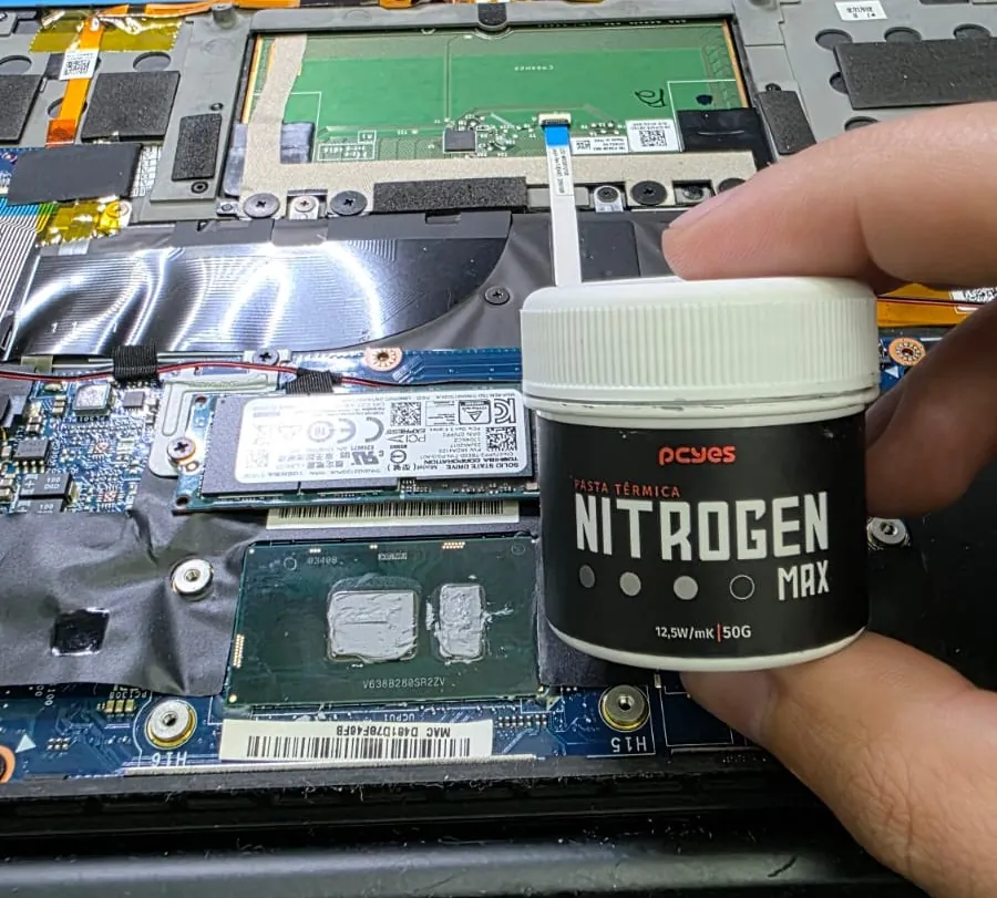 Mão segurando um pote de pasta térmica de alta condutividade sobre o interior aberto de um Dell XPS 13 9360, com a pasta antiga ainda sobre o processador