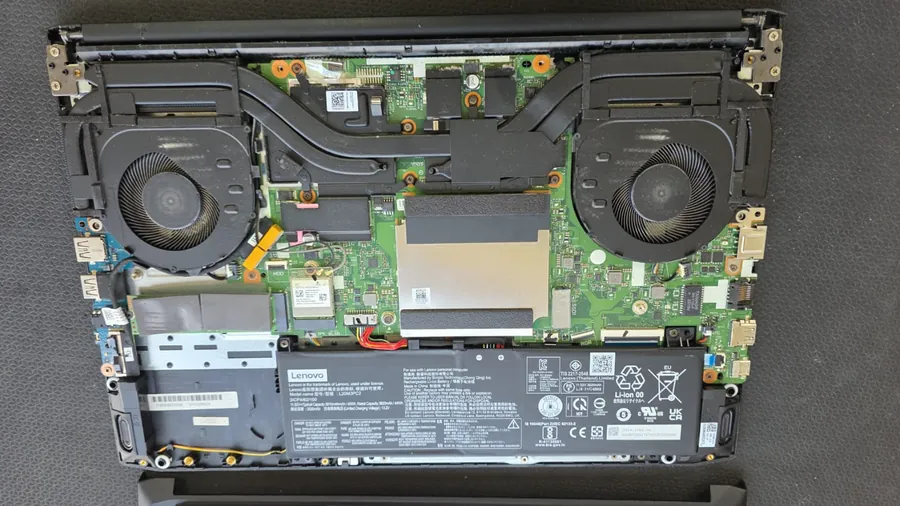 Interior de um Lenovo IdeaPad Gaming 3 com a base aberta e uma tampa metálica com espuma cobrindo os slots de memória, entre as duas ventoinhas e acima da bateria