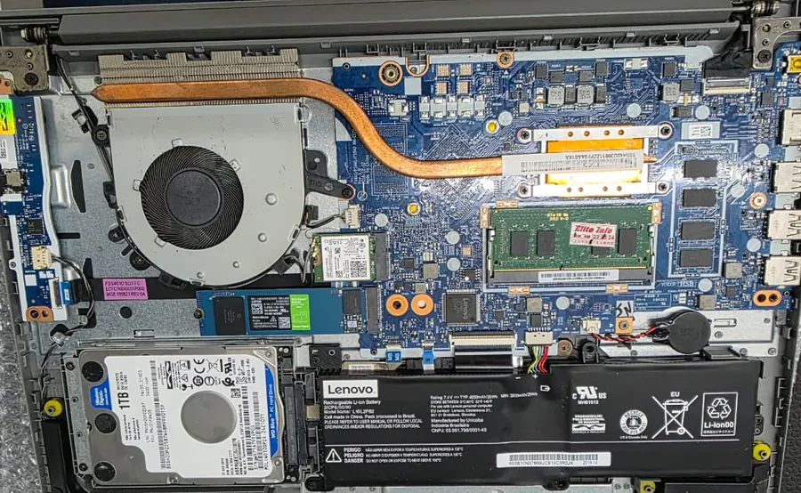 Interior de um Lenovo IdeaPad S145 com a base aberta, mostrando a ventoinha e o tubo de cobre à esquerda, o SSD M.2, o HD de 1 TB, a memória e a bateria na parte inferior